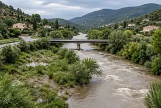 Cours d’eau en crue traversant une vallée méditerranéenne urbanisée avec berges végétalisées, pont et collines sous un ciel couvert.
